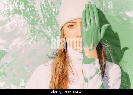 Junge Frau, die vor dem Malen steht und mit grüner Hand das Auge bedeckt Stockfoto