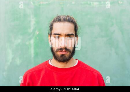 Porträt eines bärtigen jungen Mannes, der vor der grünen Wand rotes Sweatshirt trägt Stockfoto