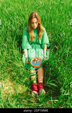 Junge Frau trägt grünes Kleid sitzt auf einem Feld suchen Am Spiegel Stockfoto