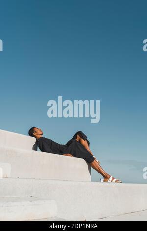 Junger Mann mit schwarzem Kaftan, der auf Betontreppen unter blauem Himmel liegt Stockfoto