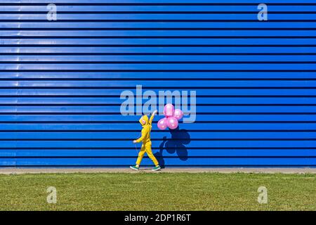 Kleines Mädchen mit Kopfhörern trägt gelben Trainingsanzug zu Fuß vor Von blauem Hintergrund mit rosa Ballons Stockfoto