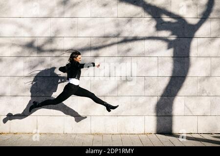 Junge Frau, die einen grossen Sprung auf eine Mauer mit Schatten eines Baumes Stockfoto