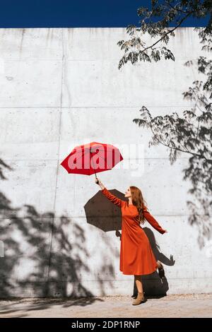 Junge Frau in rotem Kleid hält fliegenden Regenschirm während des Stehens Gegen Baumschattenwand Stockfoto