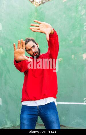 Porträt eines ernsthaften jungen Mannes mit rotem Sweatshirt, das die Hände vor der grünen Wand hebt Stockfoto