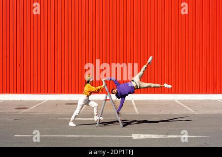 Junge Männer und Frauen, die mit einer Leiter vor sich auftreten Einer roten Wand Stockfoto