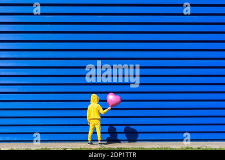 Rückansicht des kleinen Mädchens mit rosa Ballon trägt gelb tracksuit steht vor blauem Hintergrund Stockfoto