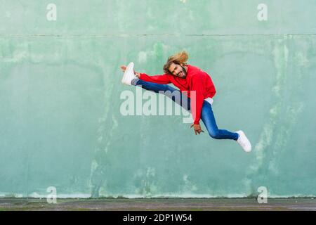 Porträt des jungen Mannes mit rotem Sweatshirt, das vor der grünen Wand in die Luft springt Stockfoto