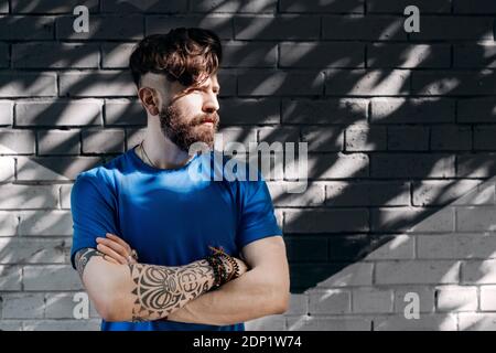 Junger Mann mit Tätowierung, der vor einer Ziegelwand steht Mit gekreuzten Armen Stockfoto