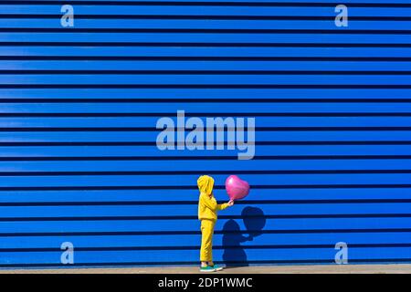 Kleines Mädchen trägt gelben Trainingsanzug stehen mit rosa Ballon in Vorderseite mit blauem Hintergrund Stockfoto