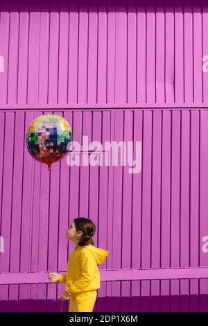 Kleines Mädchen in gelb gekleidet hält Ballon vor Violetter Hintergrund Stockfoto