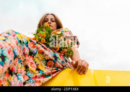 Frau im Blumenkleid sitzt am Rand des gelben Behälter Mit Blumenstrauß in ihrem Schoß Stockfoto