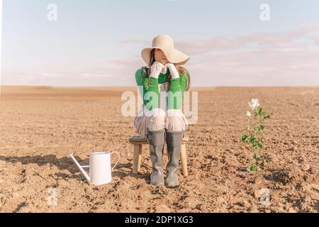 Junge Frau, die auf einem trockenen Feld neben der blühenden Pflanze auf einem Hocker sitzt Stockfoto