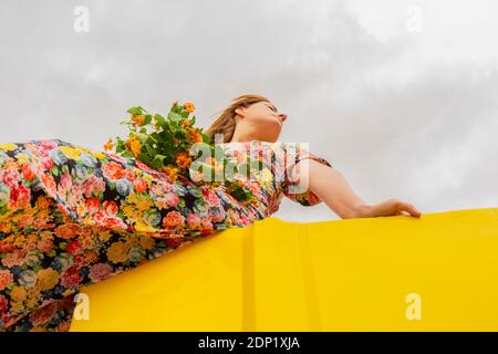 Frau im Blumenkleid sitzt am Rand des gelben Behälter Mit Blumenstrauß in ihrem Schoß Stockfoto