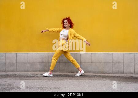 Energiegeladene junge Frau in gelbem Anzug läuft und springt hinein Vorderseite der gelben Wand Stockfoto