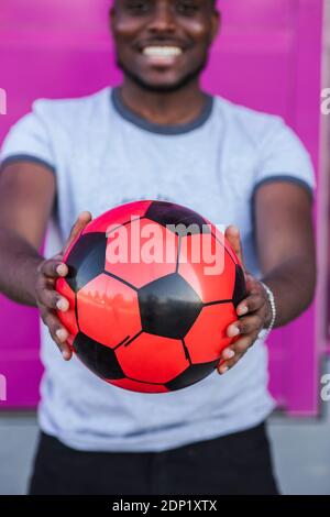 Nahaufnahme eines jungen Mannes, der den Fußball hält, während er dagegen steht Rosa Farbe Stockfoto