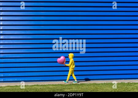 Kleines Mädchen trägt gelben Trainingsanzug zu Fuß mit rosa Ballon in Vorderseite mit blauem Hintergrund Stockfoto