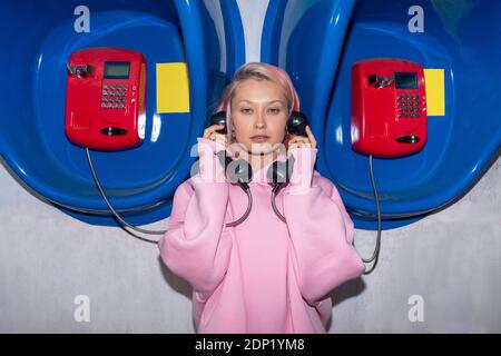 Junge Frau mit rosa Haaren trägt rosa Kapuzenhemd steht vor Telefonzellen, hält Empfänger Stockfoto