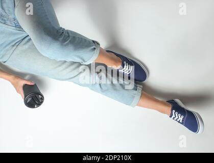 Das Mädchen in Jeans und Sneakers sitzt auf weißem Hintergrund und hält eine Tasse Kaffee. Draufsicht. Stockfoto