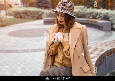 Pariser Mädchen in beigem Mantel und karierten panama Hut sitzend auf abgerundeten Bank warten auf ihr Date oder Freundinnen. Herbstspaziergang. Retro getöntes Foto Stockfoto