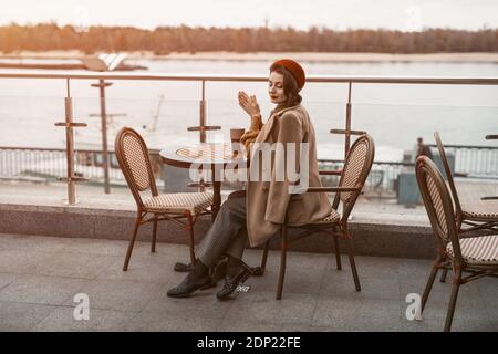 Gesamtplan einer nachdenklichen jungen Pariser Frau mit einem Kaffeebecher auf dem Tisch in roter Baskenmütze auf der Terrasse des Restaurants oder Cafés sitzen. Herbst Stockfoto