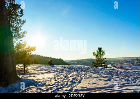 Junger Mann, der am Rand einer schneebedeckten Klippe sitzt und bei Sonnenuntergang den Blick auf den gefrorenen Fluss und das Dorf im Winter genießt. Winterlandschaft, Pine tre Stockfoto