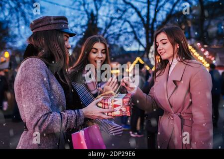 Neujahr, Weihnachtsgeschenke. Freundinnen schenken ihrem Freund auf der freien Natur eine Geschenkbox. Mädchen öffnet es. Wir feiern Winterferien unter Illuminat Stockfoto