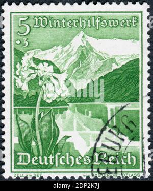 DEUTSCHLAND - UM 1938: Briefmarke gedruckt in Deutschland, Ausgabe Winterrelief, zeigt eine Zell am See (Österreich) und Bergkuhslip Blume, um 1938 Stockfoto