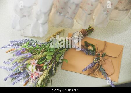 brautstrauß von Lavendel im Boho-Stil liegt auf dem Fenster neben dem handgefertigten Umschlag, auf dem Hochzeitskleid Stockfoto