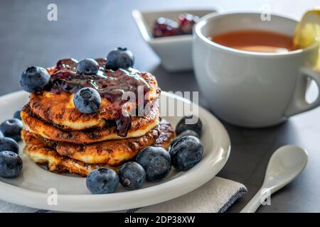 Gebratene heiße leckere Fritten von Quark mit Marmelade und Heidelbeeren auf der weißen Schale mit einer Tasse Tee auf dem Tisch. Teezeit. Stockfoto