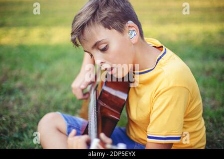 Schwerhörig preteen Junge Gitarre spielen und singen. Kind mit Hörgeräten in den Ohren, das im Park Musik spielt und singt. Hobby Kunst Aktivität für Stockfoto