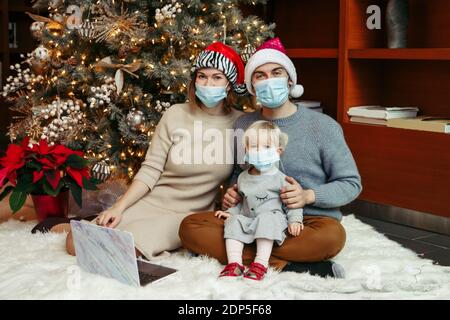 Mutter und Vater in Santa Hüte und Gesichtsmasken mit Baby Mädchen rufen Familie auf Video-Anruf. Fernere Feiertagsfeier online im Internet. Stockfoto