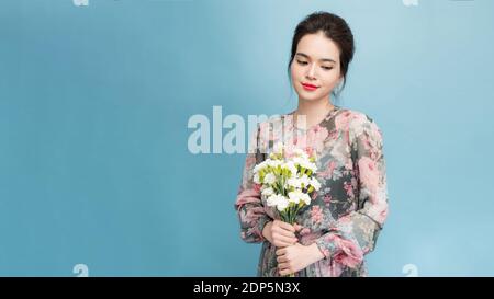 Headshot von hübschen jungen Frau mit zartem Lächeln, gesunde Haut, trägt Strauß von Frühlingsblumen, isoliert auf hellblauem Hintergrund Stockfoto