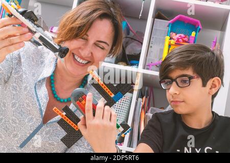 Stilvolle, kaukasische Mutter genießt das Spielen mit ihrem Sohn. Sie fliegen kleine Flugzeuge aus Plastikziegeln. Frau lacht. Moderne Familienaktivitäten mit Kindern. Stockfoto