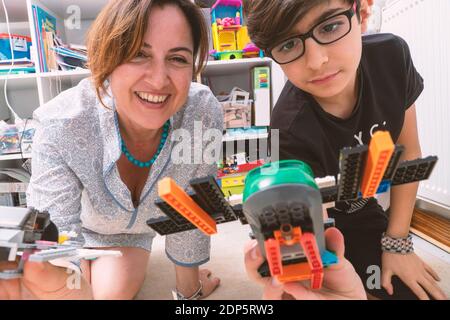 Fröhliche Mutter und ihr Sohn spielen mit Block Spielzeug Flugzeuge. Sie sitzen auf dem Boden im Kinderzimmer vor einem Regal. Frau lächelt Stockfoto