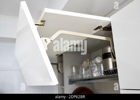 Teil des modernen Kücheninterieurs. Offener weißer horizontaler Küchenschrank mit Tellern und Gläsern auf Regal. Tür des Küchenschranks anheben Stockfoto