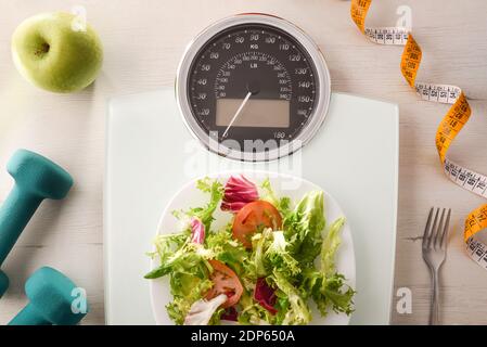 Gewichtskontrollkonzept mit Waage mit Salatplatte und Besteck auf dem Tisch und Obst, Gemüse und Sportzubehör auf dem Tisch. Draufsicht. Horizontal Stockfoto