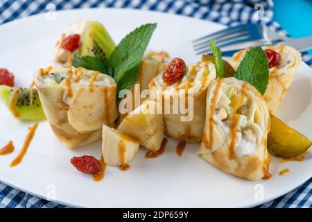 Einfache hausgemachte Pfannkuchen mit Banane, Kiwi, Erdbeermarmelade auf einem weißen Teller geschnitten. Cremige Quark-Füllung. Das Essen ist mit Minze dekoriert. Dünn gerollt Stockfoto