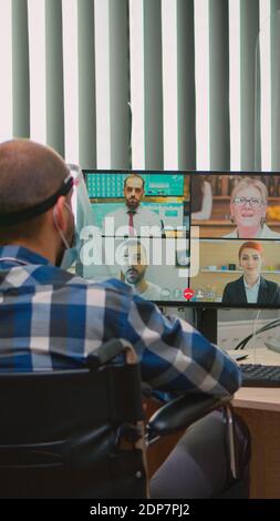 Gelähmter Manager im Rollstuhl im Gespräch mit Maske und Visier während Videocall mit Online-Konferenz im Geschäftsbüro. Immobilisierte Freiberufler arbeiten in Finanzunternehmen unter Wahrung der sozialen Distanz Stockfoto