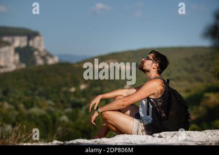 Junger männlicher Wanderer mit Rucksack, der sich während des ruhigen Sonnenuntergangs auf einem Berg entspannt - Landschaft aus dem Urlaub. Müder Reisender Mann, der auf einem Hügel ruht. Stockfoto