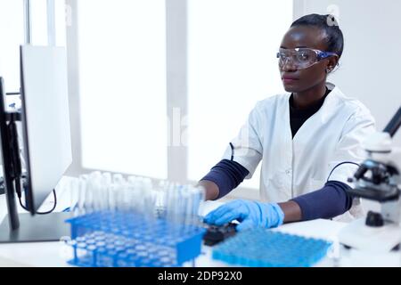 Afrikanischer Biotechnologieforscher arbeitet im hellen modernen Labor mit Computer. Multiethnischer Gesundheitswissenschaftler in biochemischen Einrichtungen, der sterile Geräte trägt. Stockfoto