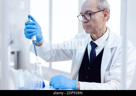 Älterer Wissenschaftler, der pharmazeutische Experimente mit Pipette macht. Leitender professioneller Chemiker, der Pippete mit blauer Lösung für mikrobiologische Tests verwendet. Stockfoto