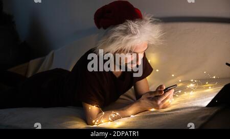 Hübscher junger Mann, der auf der Couch liegt und einen Weihnachtsmütze trägt, sein Telefon benutzt und in einem dunklen weihnachtlich dekorierten Zimmer lächelt. Es ist angenehm, Zeit zu verbringen Stockfoto