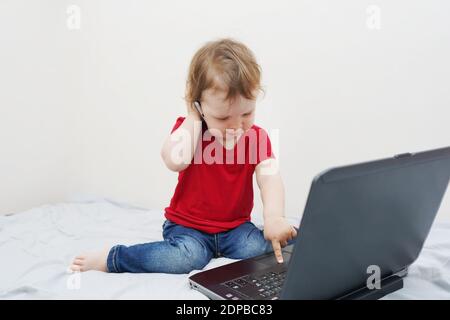 Nettes kleines Mädchen sitzt auf dem Bett mit ihrem Laptop Und Telefon in der Nähe des Ohrs halten Stockfoto