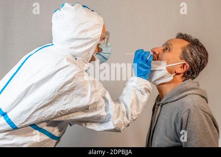 Ein Arzt, der vollständig mit einem weißen Overall, einer Schutzbrille, einer Maske und Handschuhen bedeckt ist, führt einem Patienten mittleren Alters einen Nasenabstrich für eine covid-19 che durch Stockfoto