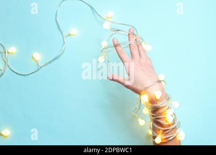 Minimalismus-Trend. Weibliche Hand mit hellen leuchtenden Girlanden von Glühbirnen auf blauem Hintergrund gewickelt. Draufsicht Stockfoto