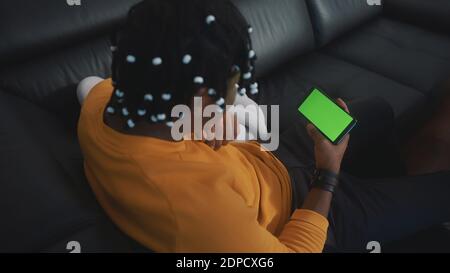 Smartphone mit grünem Bildschirm. afroamerikanischer schwarzer Mann mit geflochtenen Haaren, der sich auf der Couch entspannt und auf den Telefonbildschirm schaut. Hochwertige Fotos Stockfoto
