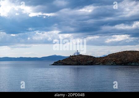 Griechenland, Insel Kea Tzia. Weißer Leuchtturm auf einem felsigen Kap, wolkiger blauer Himmel und welliger dunkler farbenfroher Meeresgrund. Sommer, Touristenziel Stockfoto
