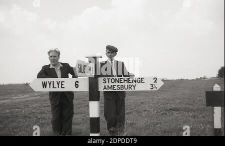 1950er Jahre, historisch, auf der Salisbury Plain, zwei Männer, einer trägt ein Tuch oder eine flache Kappe, typische männliche Kopfbedeckung aus der Zeit, stehen an Straßenschildern, einer weist auf Stonehenge, das berühmte prähistorische Denkmal der alten stehenden Steine, die auf der Ebene liegen, in Wiltshire, England, Großbritannien. Stockfoto