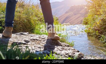 Tourist in Jeans und Turnschuhen Spaziergänge entlang der Ufer des Bergflusses zwischen Felsen und grünen Pflanzen und Stufen über Bach an sonnigen Tag aus nächster Nähe. Stockfoto