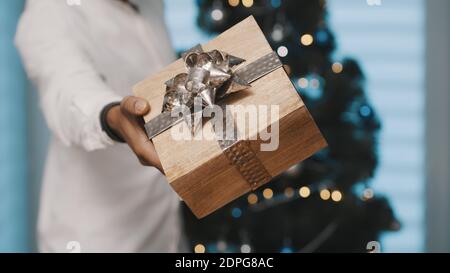 afroamerikanischer schwarzer Mann, der Weihnachtsgeschenk mit Goldband vor dem weihnachtsbaum gibt. Nahaufnahme. Hochwertige Fotos Stockfoto
